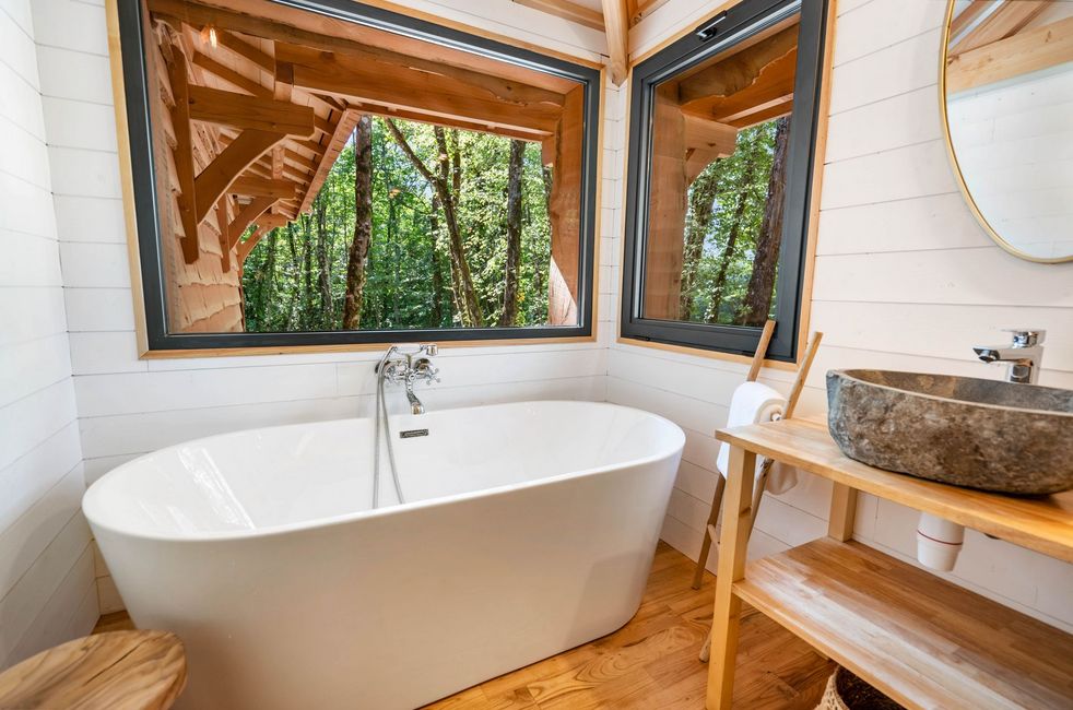 Salle de bain du lodge Kali de Calviac avec deux lavabos, deux miroirs, une baignoire et une fenêtre avec vu sur les bois